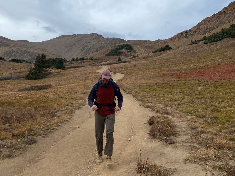 autumn high alpine mountain range with red, beige, and green grass. Path going up towardds mountains with man walking towards camera looking at ground.