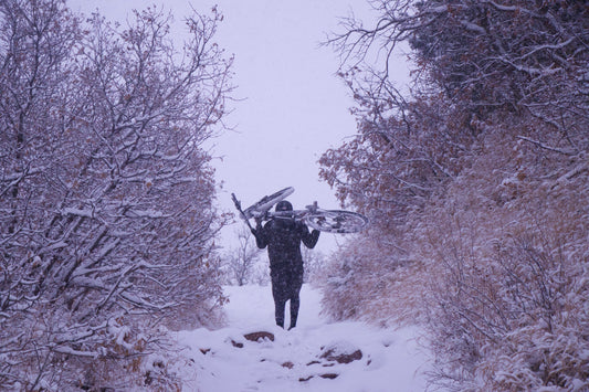 image of a man walking up a snowy trail in a blizzard with a bike over his shoulders and shrubs on either side of the trail.